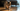 A domestic short-haired cat crouching on a hardwood floor, mouth slightly open as if gagging